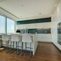 Interior kitchen; all-white walls, cabinets, ceiling, island and three chairs; silver appliances on right; large windows on left