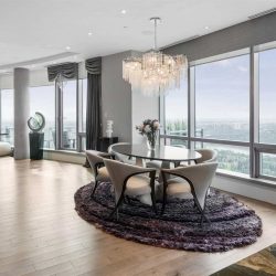 Interior penthouse rounded living room with hardwood floor and large windows; small black dining table with six white cloth chairs around it, on top of purple shag carpet, white chandelier overhead; white ceiling, light grey walls and curtain