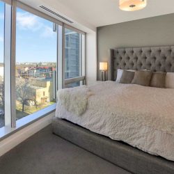 Interior condo bedroom, light grey floor, white ceiling and wall; grey frame bed with white comforter; large window to left