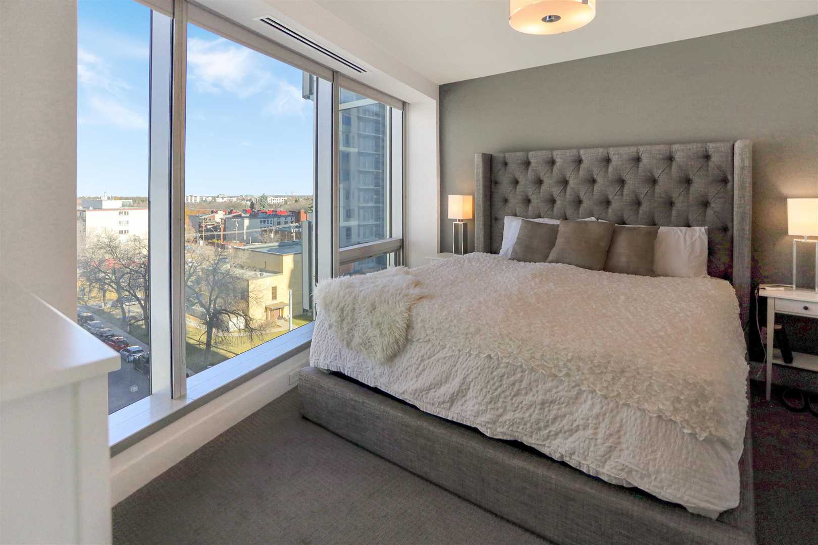Interior condo bedroom, light grey floor, white ceiling and wall; grey frame bed with white comforter; large window to left