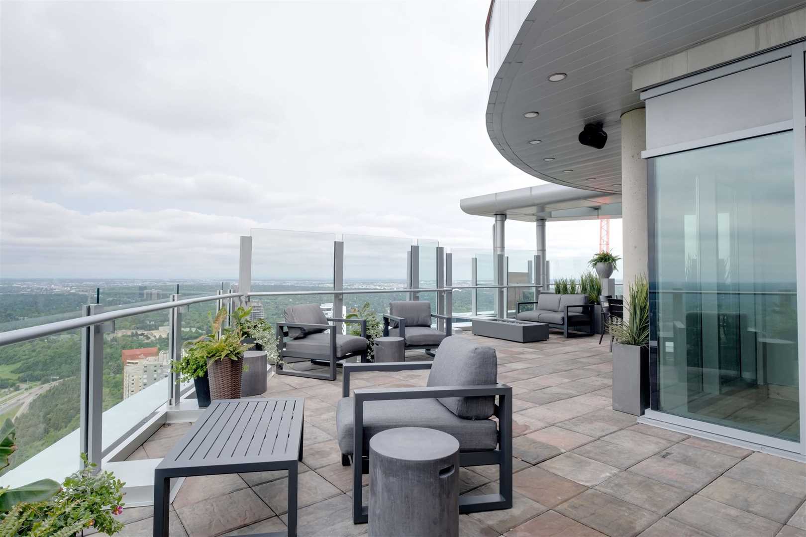 Penthouse curved patio with light grey and red stone floor on left, white building on right; dark grey cloth chairs and green potted plants throughout