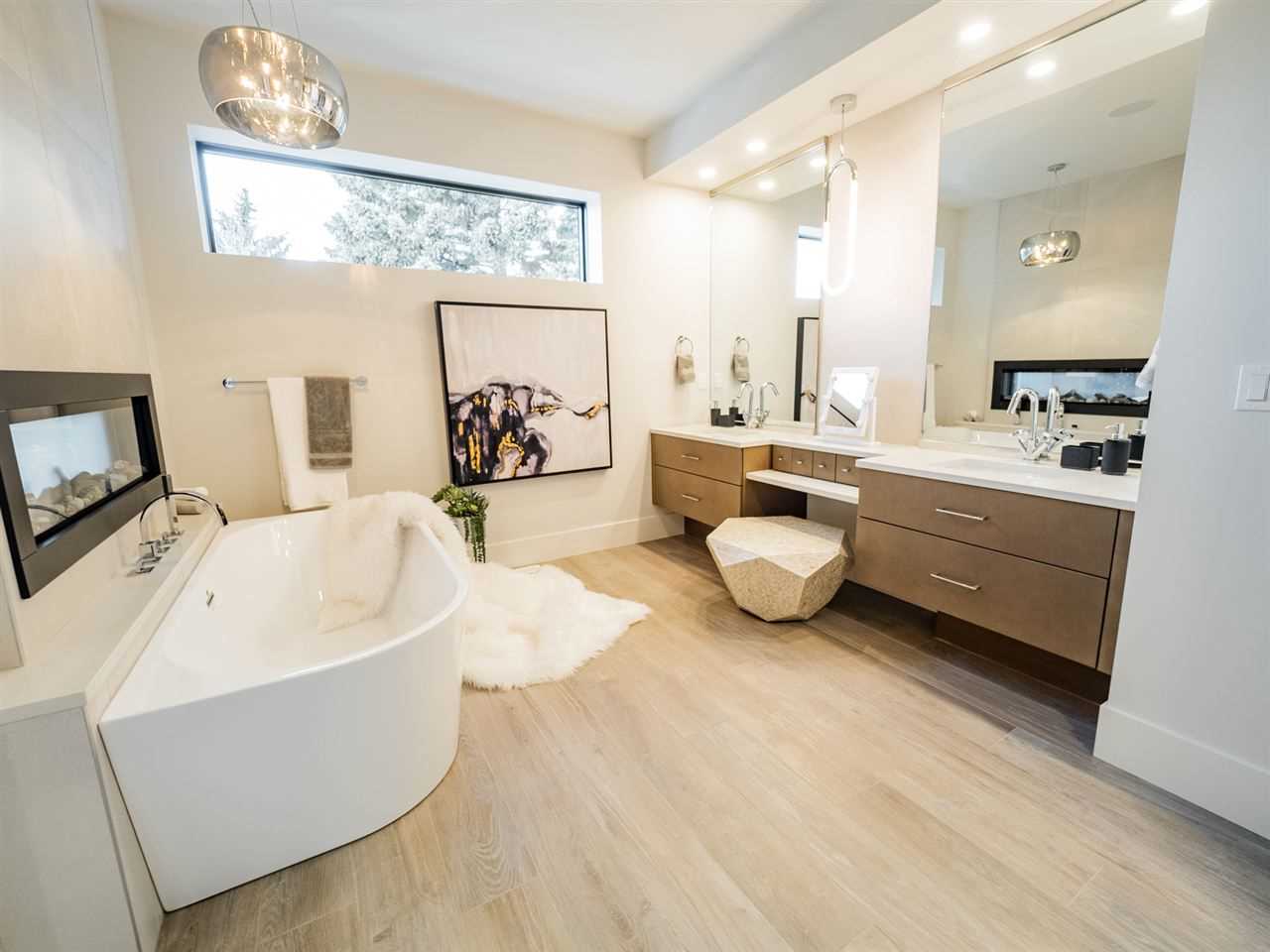 Interior en suite bathroom with white oak floor, white ceiling and walls; his-and-her sinks above brown wood drawers on the right; rectangular window at head height on back wall; white soaker tub against two-way fireplace on the left