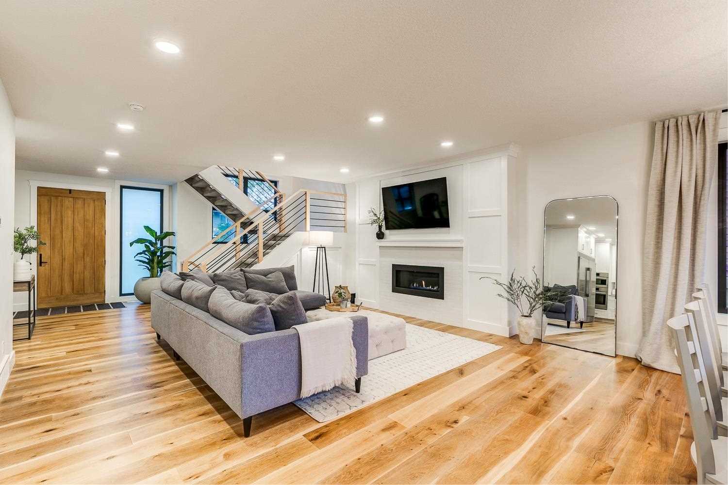 Interior living room, white ceiling and walls, light hardwood floor; grey sectional couch facing right to TV mounted above fireplace; open-air stairs background to the left; large mirror against the wall to the right