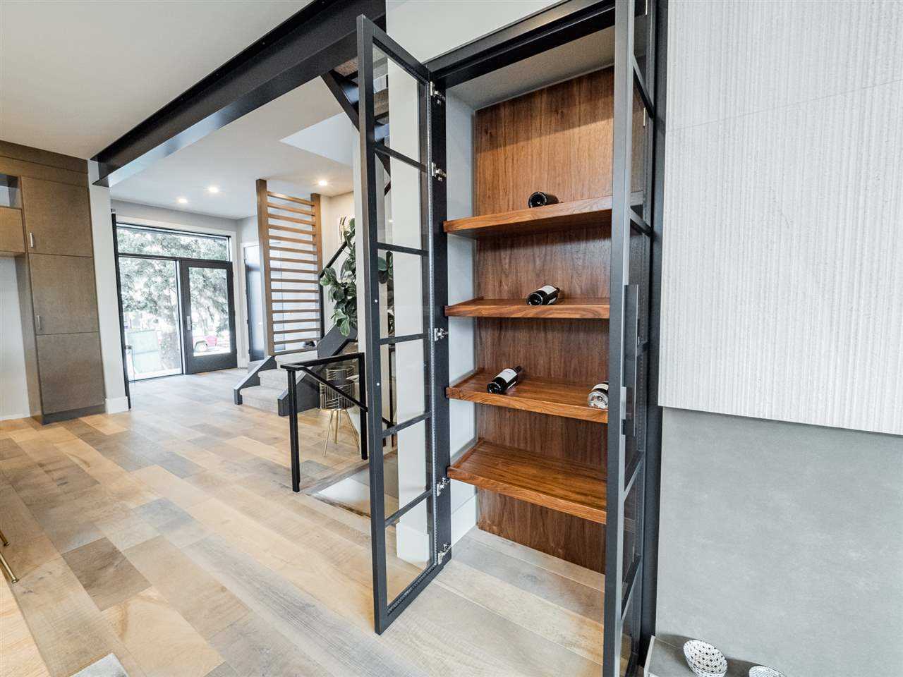 Interior shot of wine rack with entrance to the left in the background; black-framed glass doors open to mostly empty four-shelf wood wine rack; white oak floors lead to entrance; black beam on white ceiling