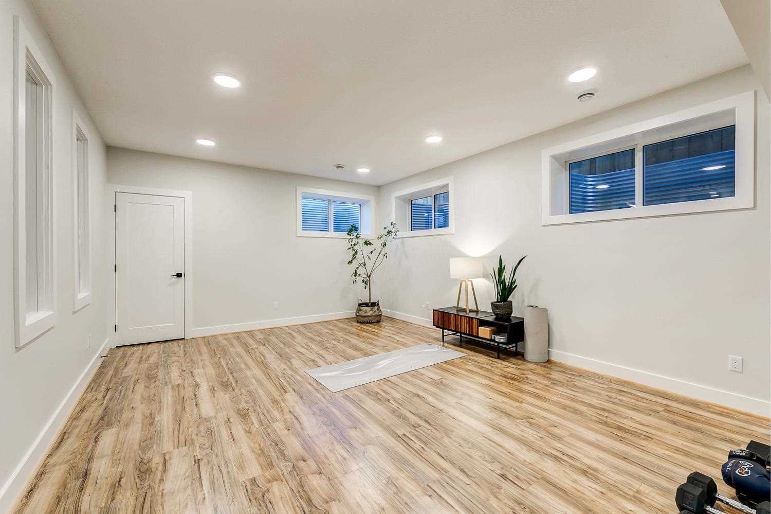 Basement yoga studio with white ceiling and walls, light hardwood floor and a white yoga mat; small table against wall with lamp and plant on top; three ground-level windows on the middle and right; plant in the corner