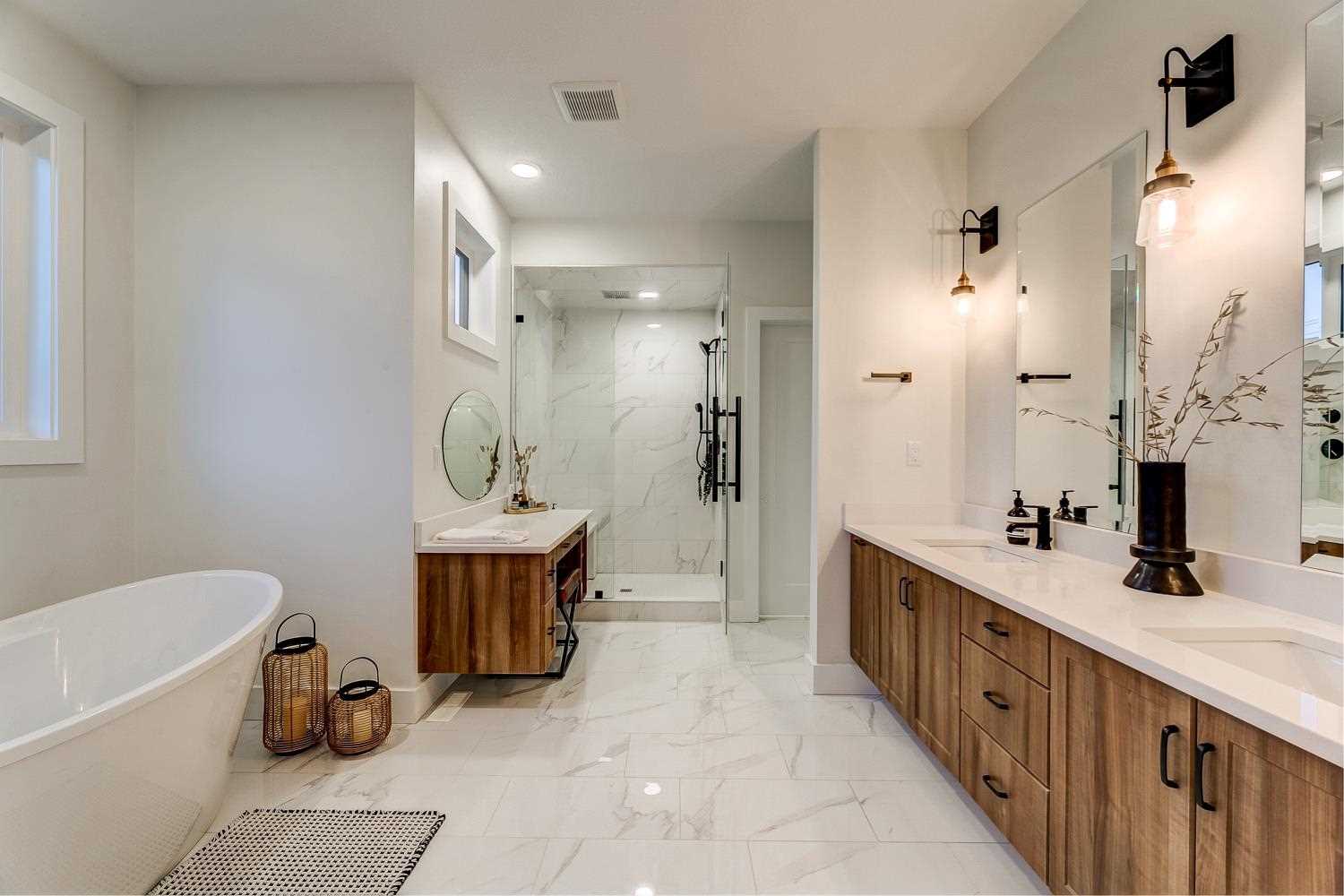 En suite bathroom with white ceiling and walls, light hardwood floor; wood cupboards and large mirror on right; glass shower in background with wood makeup counter with round mirror in front; white soaker tub to the left