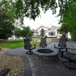 Backyard view from dark grey stone fire pit with four chairs surrounding it and trees on either side; lawn leading to deck and house