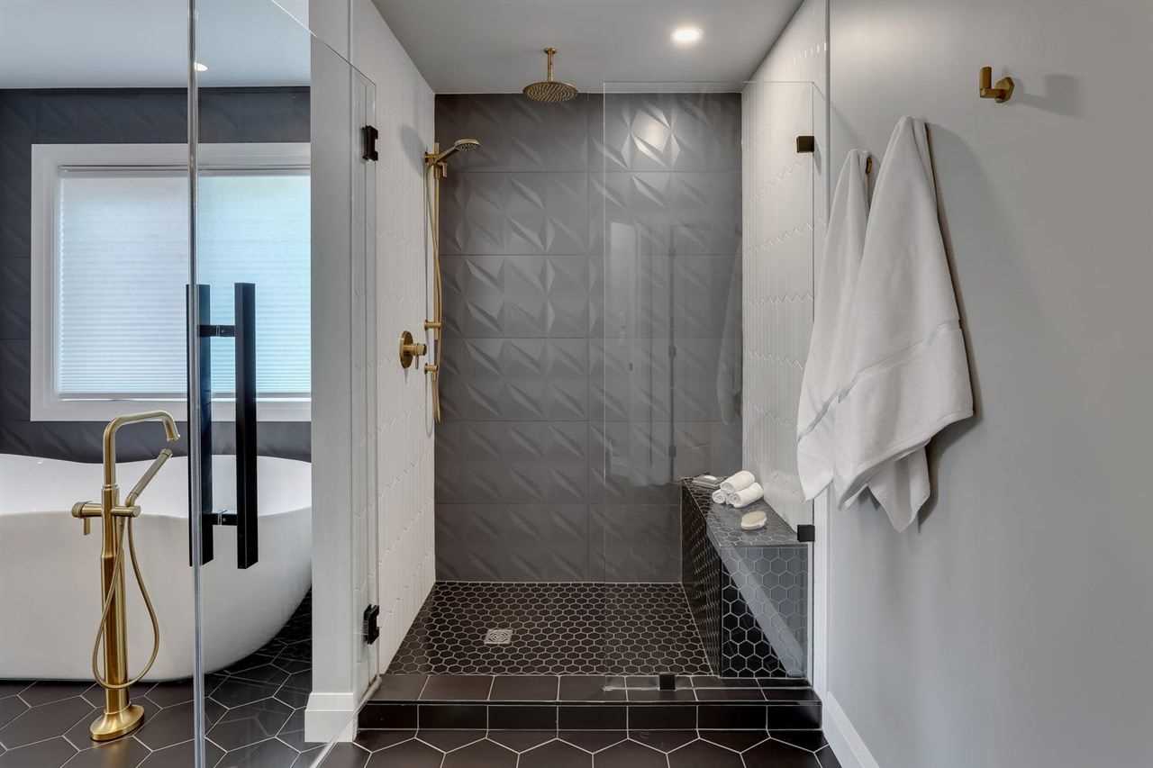 En suite bathroom, white wall, black penny tile floor; glass door shower with two white walls, one grey textured wall, black penny tile floor