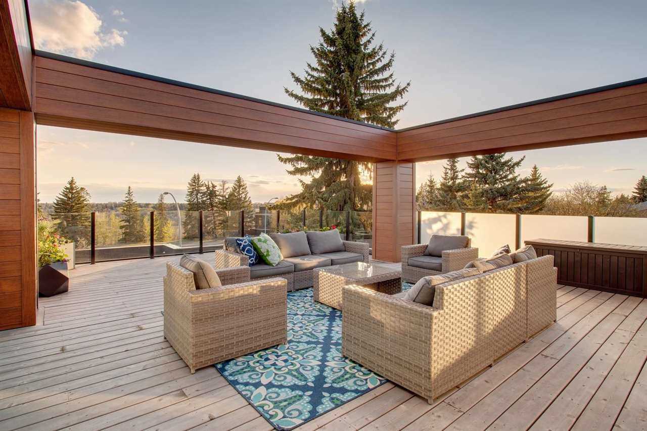 Rooftop patio with wood plank floor; beige and grey wicker couches and chairs around beige wicker coffee table; wood beams making a rectangle above, glass rail around floor; pine tree in background