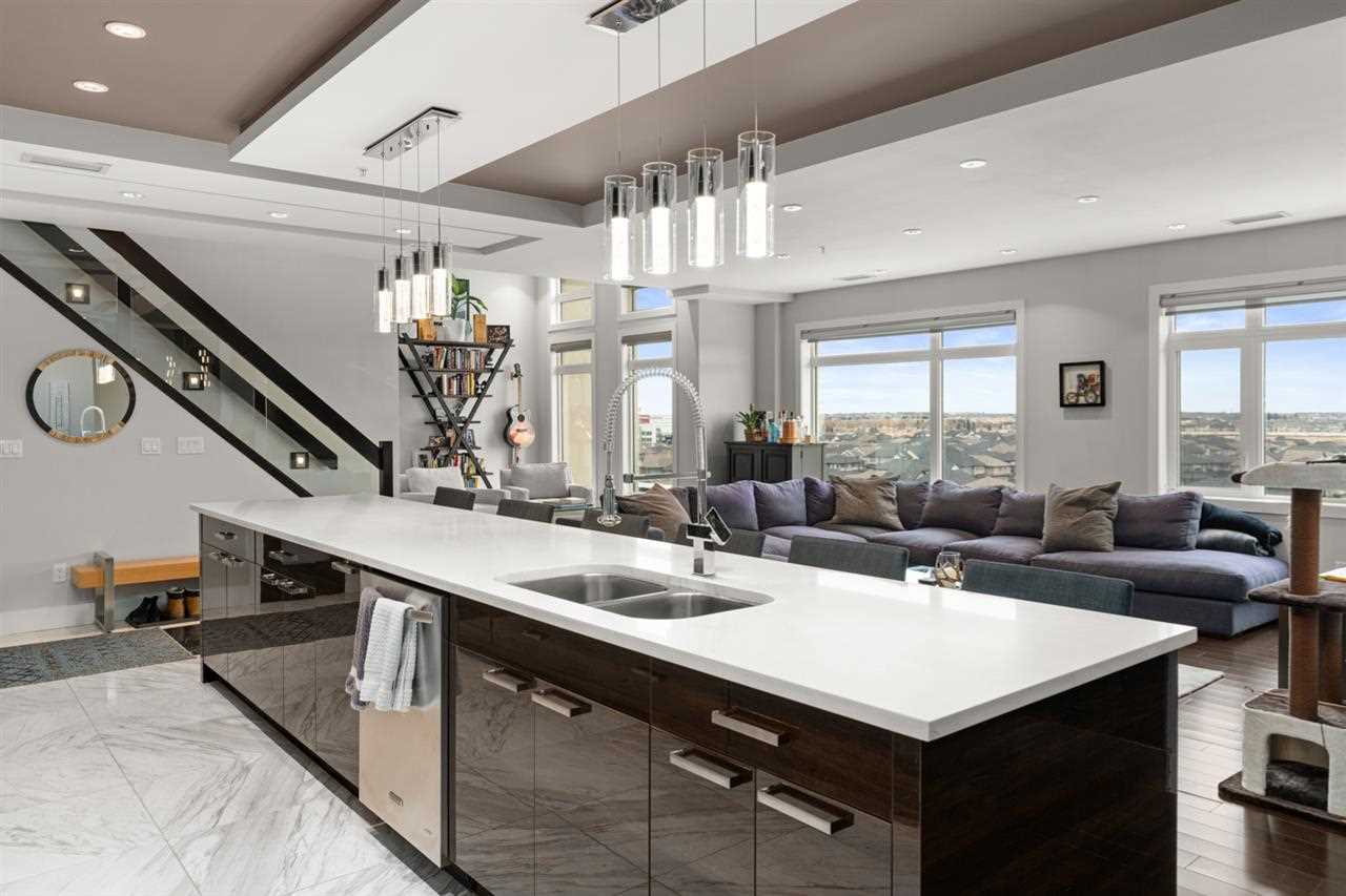 Interior kitchen-living room, white marble kitchen floor, dark hardwood living room floor; black island with white counter in foreground, two sets of four lights hanging above; large grey sectional in front of large windows in background; stairs to the left