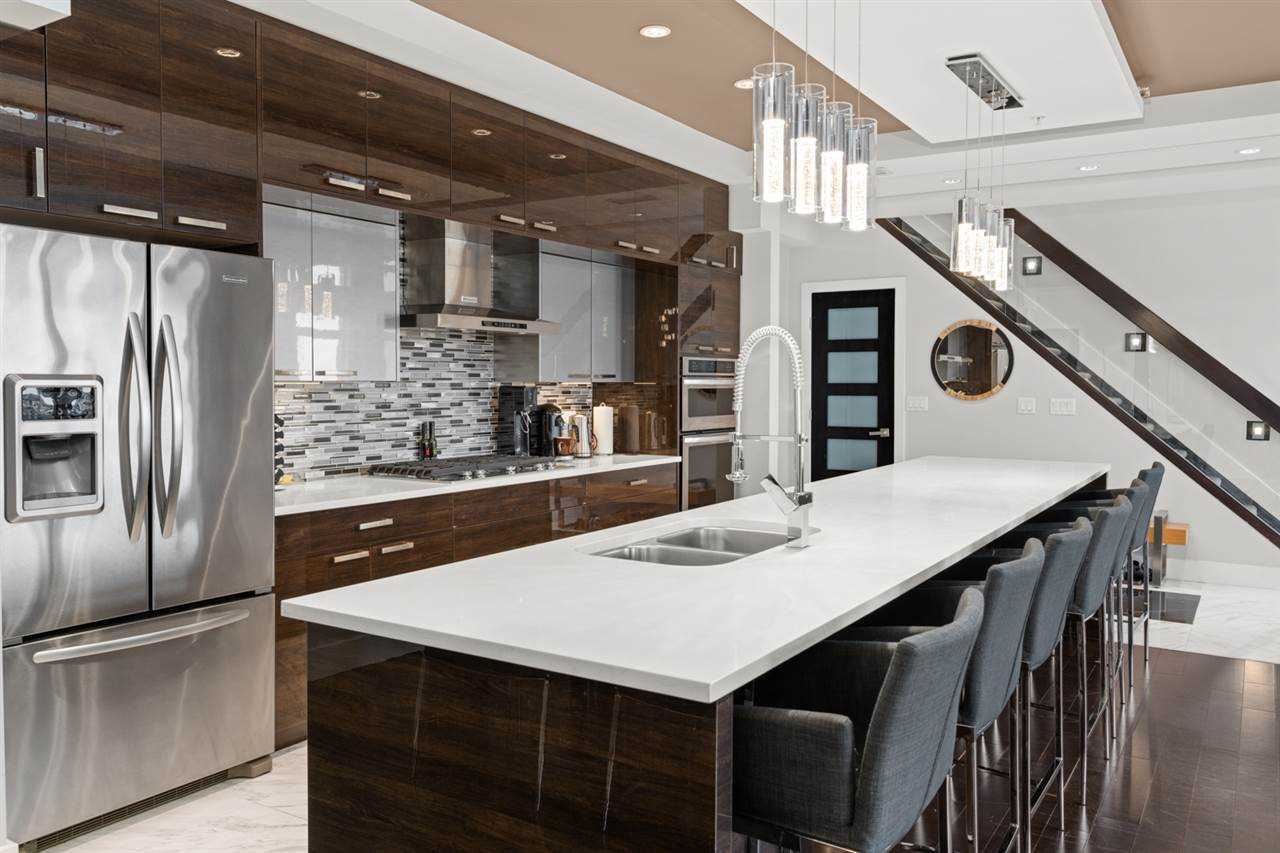 Black kitchen island with white counter, six chairs on right side, two sets of four lights hanging above; dark brown cabinet, black and white tile backsplash, silver fridge on left, stairs on right