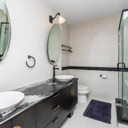 Interior en suite bathroom; white floor and walls, two sinks with single faucets and round windows above; stand-up glass shower and soaker tub on the right
