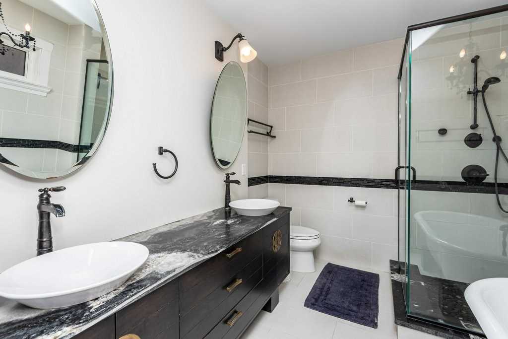 Interior en suite bathroom; white floor and walls, two sinks with single faucets and round windows above; stand-up glass shower and soaker tub on the right