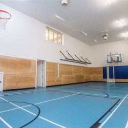 Interior basketball court with blue floor, light wood panelling on bottom half of white wall; windowed viewing area on top left