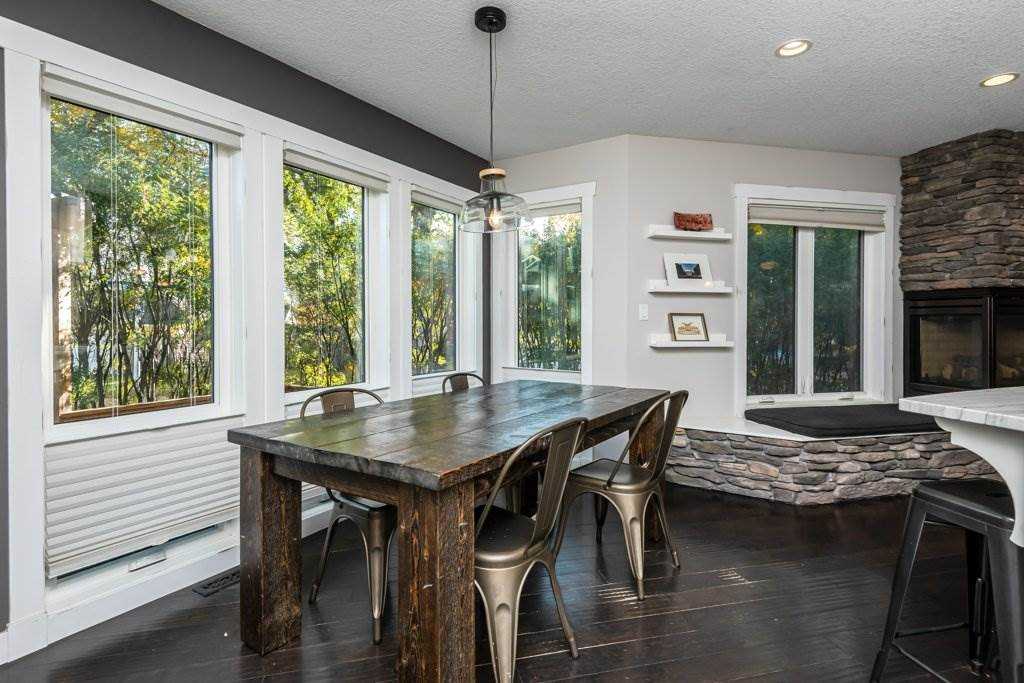 Interior dining room, dark wood floor and table, silver steel chairs, white windows and walls, hanging Edison lightbulb over the table, dark stone fireplace 