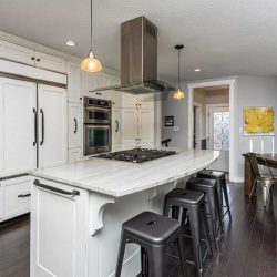 Interior kitchen, dark wood floors, white cabinets and island with silver stools around it, hood fan and two hanging lights over it