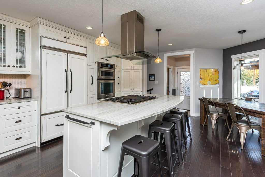 Interior kitchen, dark wood floors, white cabinets and island with silver stools around it, hood fan and two hanging lights over it