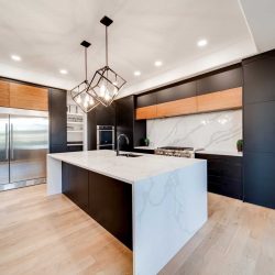 Kitchen with white ceiling and walls, light hardwood floor; black cupboards with white backsplash; white marble waterfall island with two hanging geometric lights above