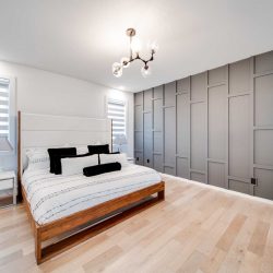 Master bedroom, white ceiling and walls, light hardwood floor; grey textured feature wall on right; geometric light above white bed with wood frame