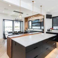Kitchen with white ceiling and walls, light hardwood floor; black cabinets and drawers, including on island with white counter and wood extension and three hanging lights above; sink to the right, with windows and small TV above; dining table beyond kitchen area