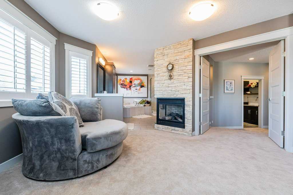 Open concept en suite with round grey lounger in front, double-sided fireplace on wall, corner jacuzzi tub in background