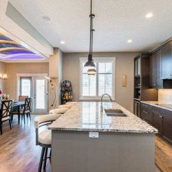 Interior kitchen, hardwood floor, white ceiling and walls, looking over white granite island with sink on right, four stools on left, three black lights hanging overhead
