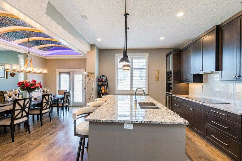 Interior kitchen, hardwood floor, white ceiling and walls, looking over white granite island with sink on right, four stools on left, three black lights hanging overhead