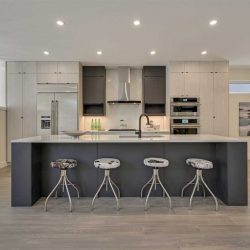 Interior kitchen with grey hardwood floor and white ceiling with embedded circular lights; black base island with white top (and sink) and room to slide four stools under its overhang; light grey cabinets with two black shelves on either side of hood fan above stove