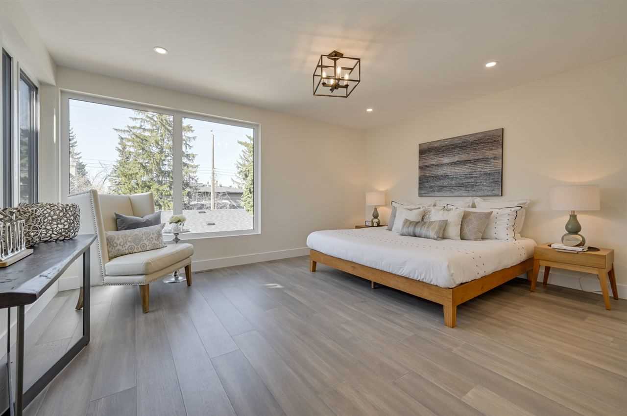 Master bedroom with grey hardwood floor, white ceiling (with small, box-framed light in middle) and walls; large bed with white covers on wood frame, wood night stand beside; white chair in front of large windows to the left