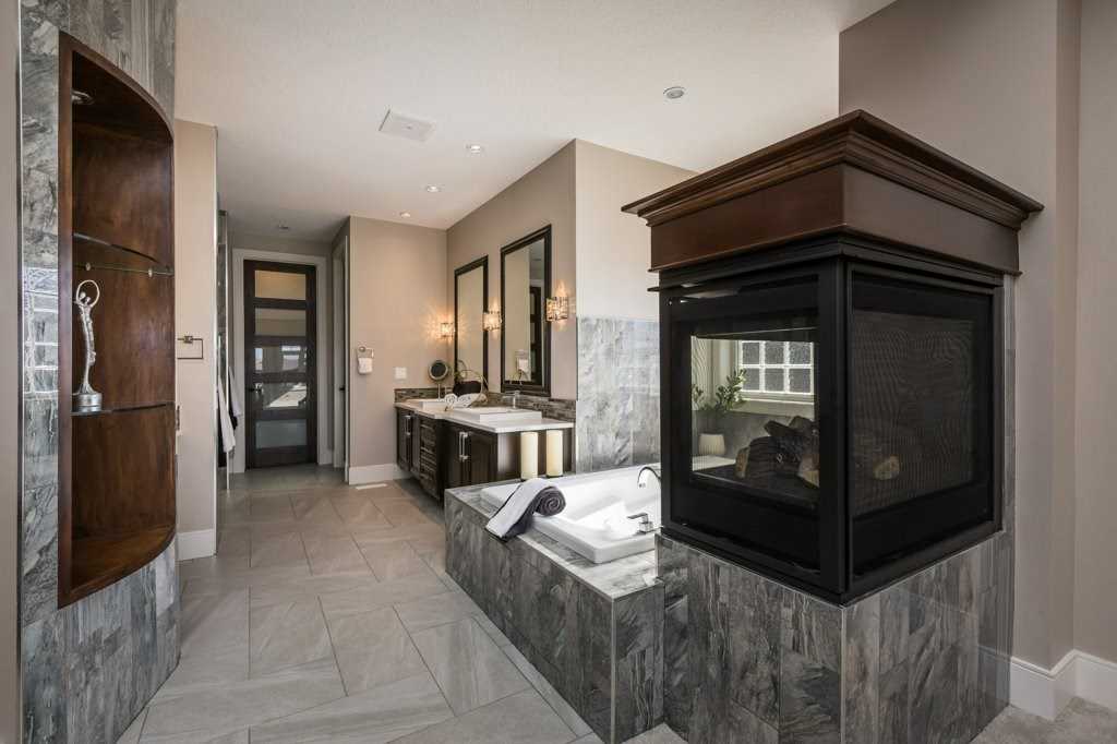 En suite bathroom with three-sided fireplace, light marble floor, wood shelving, large tub and his-and-her sinks