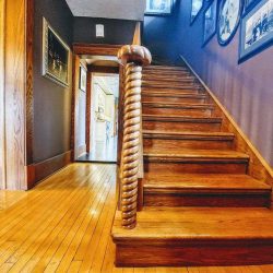 Interior entrance looking at hand-crafted wood stairs with a spiral post railing at the bottom; hardwood flooring leading to kitchen straight ahead, and to living room on the left