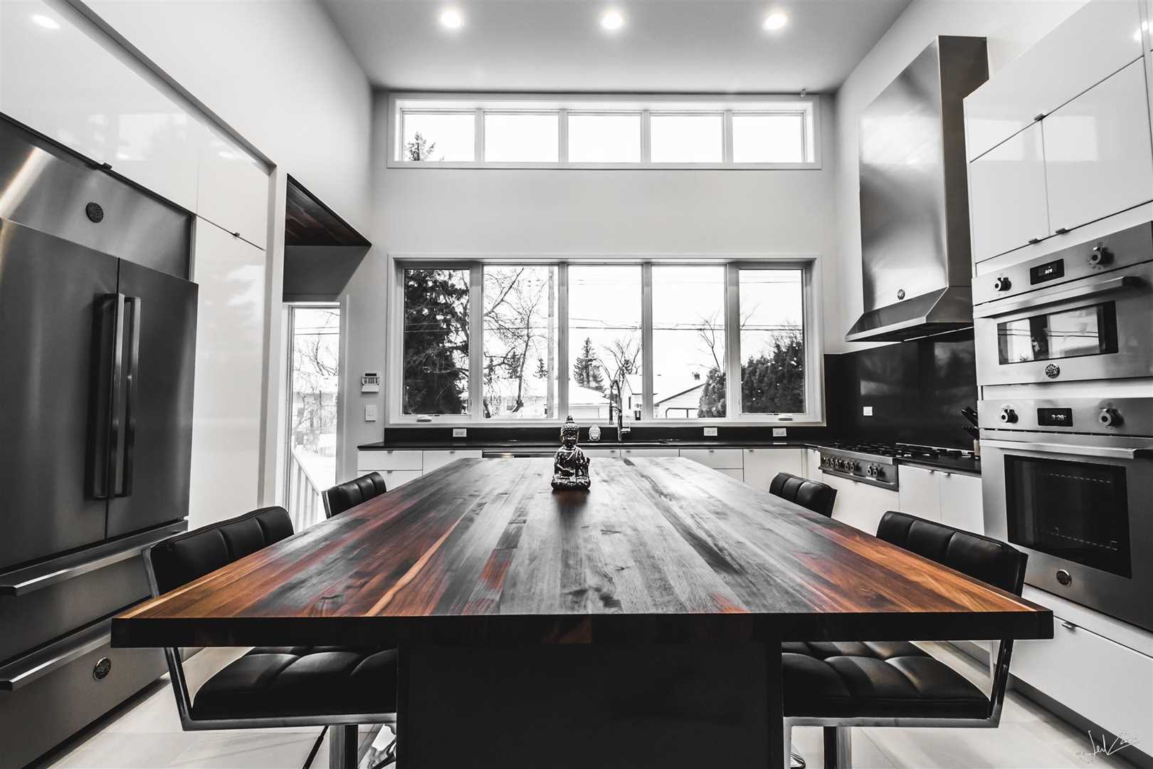 Dark wood table in centre of white kitchen with black appliances on left and right; looks out large windows in background