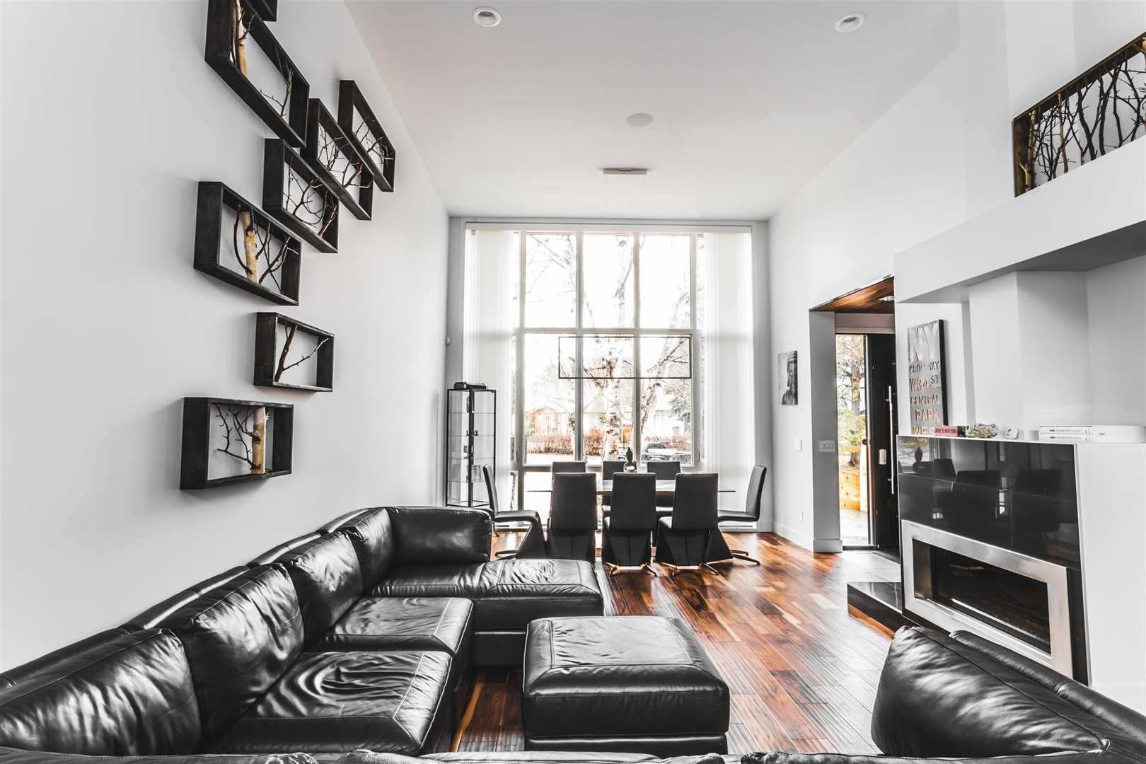 White living room facing out through floor-to-ceiling windows, hardwood floors; black sectional on left with framed tree branches above; fireplace on right