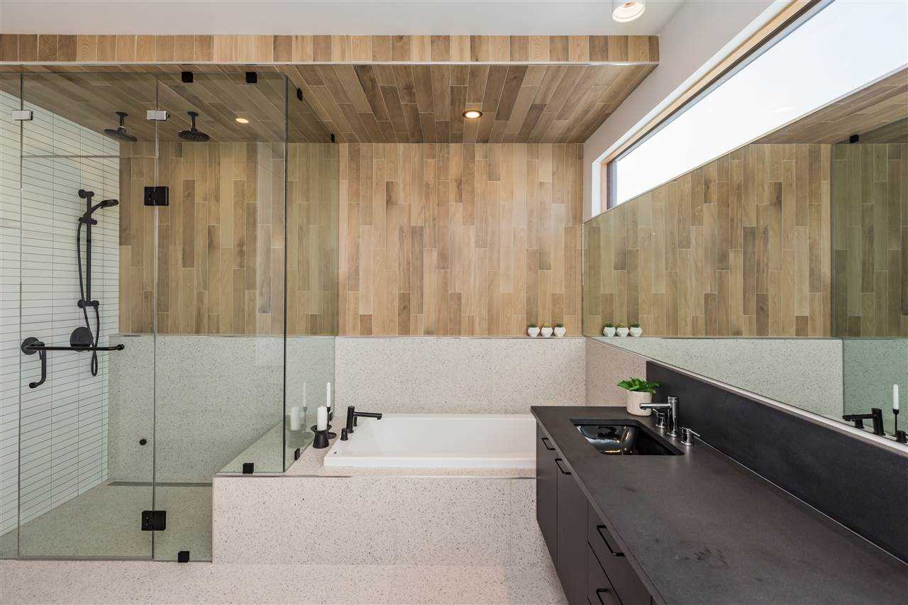 Interior en suite; glass-wall shower divided from sunk-in tub; light speckled tile floor and tub mount; light wood panel walls; black sink and full-width mirror on right