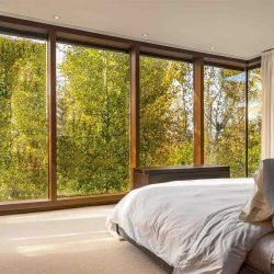 Master bedroom with white carpet and ceiling; off-white bed faces floor-to-ceiling, wood-framed windows  looking out to trees