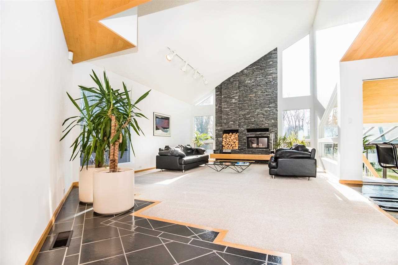 White-walled living room with light carpet and dark grey furniture. Giant windows and fireplace on triangular wall. 