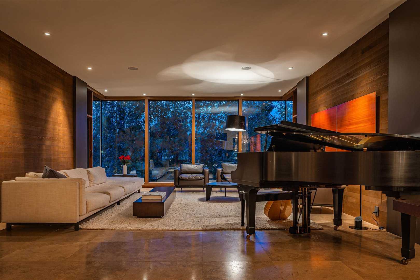 Interior living music room, brown marble floor and wood walls, white ceiling; grand piano to right with a guitar sitting on the other side; white cloth couch on left, two leather chairs centre, white shag carpet in the middle; floor-to-ceiling windows in background 