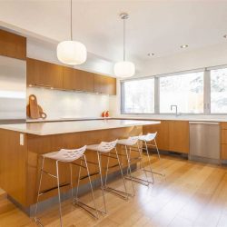 Interior kitchen, hardwood floor and cabinets, white countertops; four silver and white stools at island, two white lights overhead; windows on right