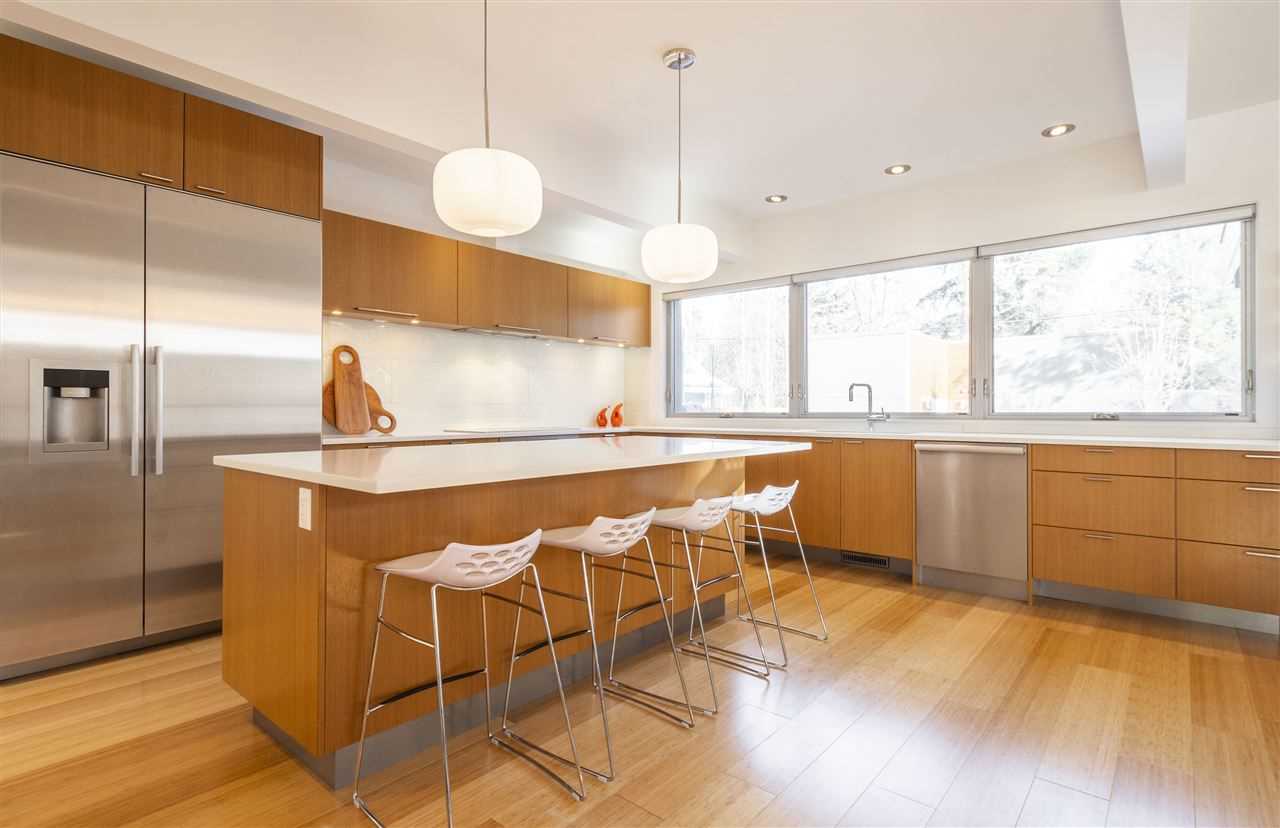 Interior kitchen, hardwood floor and cabinets, white countertops; four silver and white stools at island, two white lights overhead; windows on right