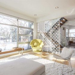 Interior all-white living room, looking out front window; white footrest and chair on top of white shag carpet on top of hardwood floor; yellow chair in middle of the shot; circular-holed divider between room and two-flight stairs