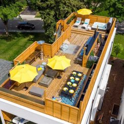 All-wood rectangular rooftop patio; black table on blue rug with eight black chairs and eight yellow dinner plates bottom right; two yellow umbrellas with grey furniture to the left; stainless steel outdoor kitchen middle-right; hot tub top right next two another yellow umbrella and two chaise loungers 
