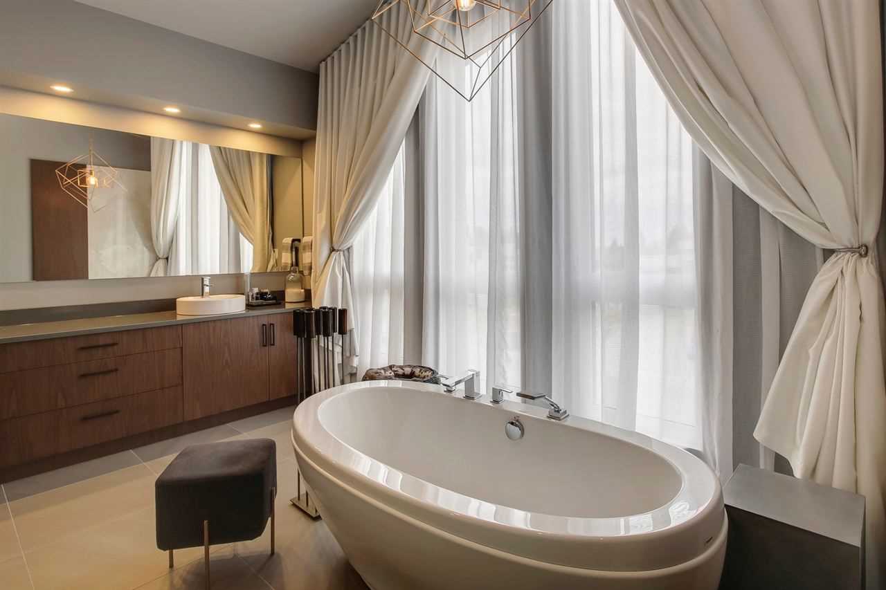 En suite bathroom with free-standing soaker tub, black stool, wood cabinets and drawers under a large mirror; curtains over the windows. 