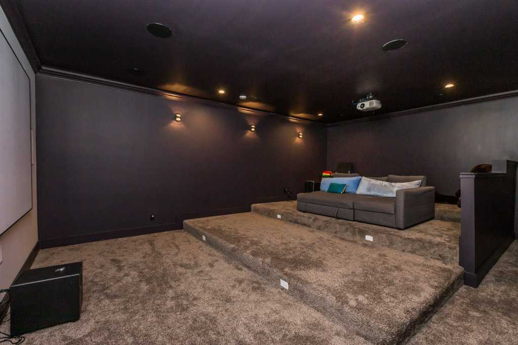 Theatre room with dark grey carpet and purple walls; projector hangs above deep couch which sits on third level of the terraced floor; large screen on the left